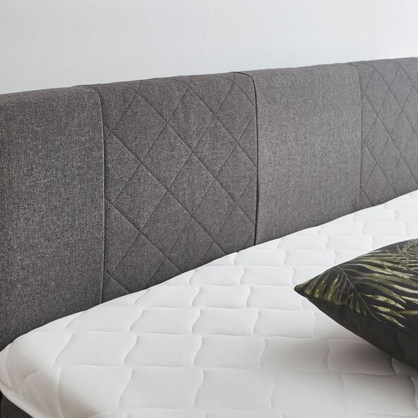 Graues Boxspringbett Basic mit großem Kopfteil, bestehend aus Matratze und Unterbau, im Schlafzimmer vor heller Wand.