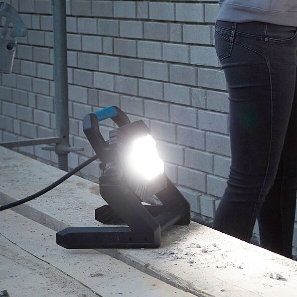 LED Arbeitsstrahler mit schwarzem Gehäuse und Griff, der auf einem kippbaren Bügel montiert ist. Das Licht scheint hell und gleichmäßig.