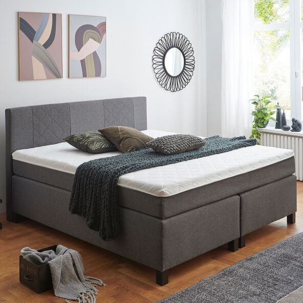 Graues Boxspringbett Basic mit großem, rechteckigem Kopfteil und strukturierter Oberfläche, im Schlafzimmer auf hellem Holzboden stehend.