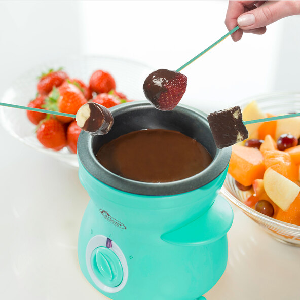 Türkisfarbenes Mini Schoko-Fondue Set mit Teelicht, Schokoladenstückchen, zwei Gabeln und einem Herz-Keks auf einem weißen Untergrund.