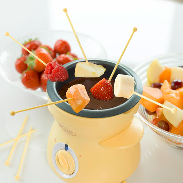 Gelbes Mini Schoko-Fondue Set mit Schokoladenstückchen und kleinen Gabeln, bereit für den Gebrauch auf einer weißen Oberfläche.