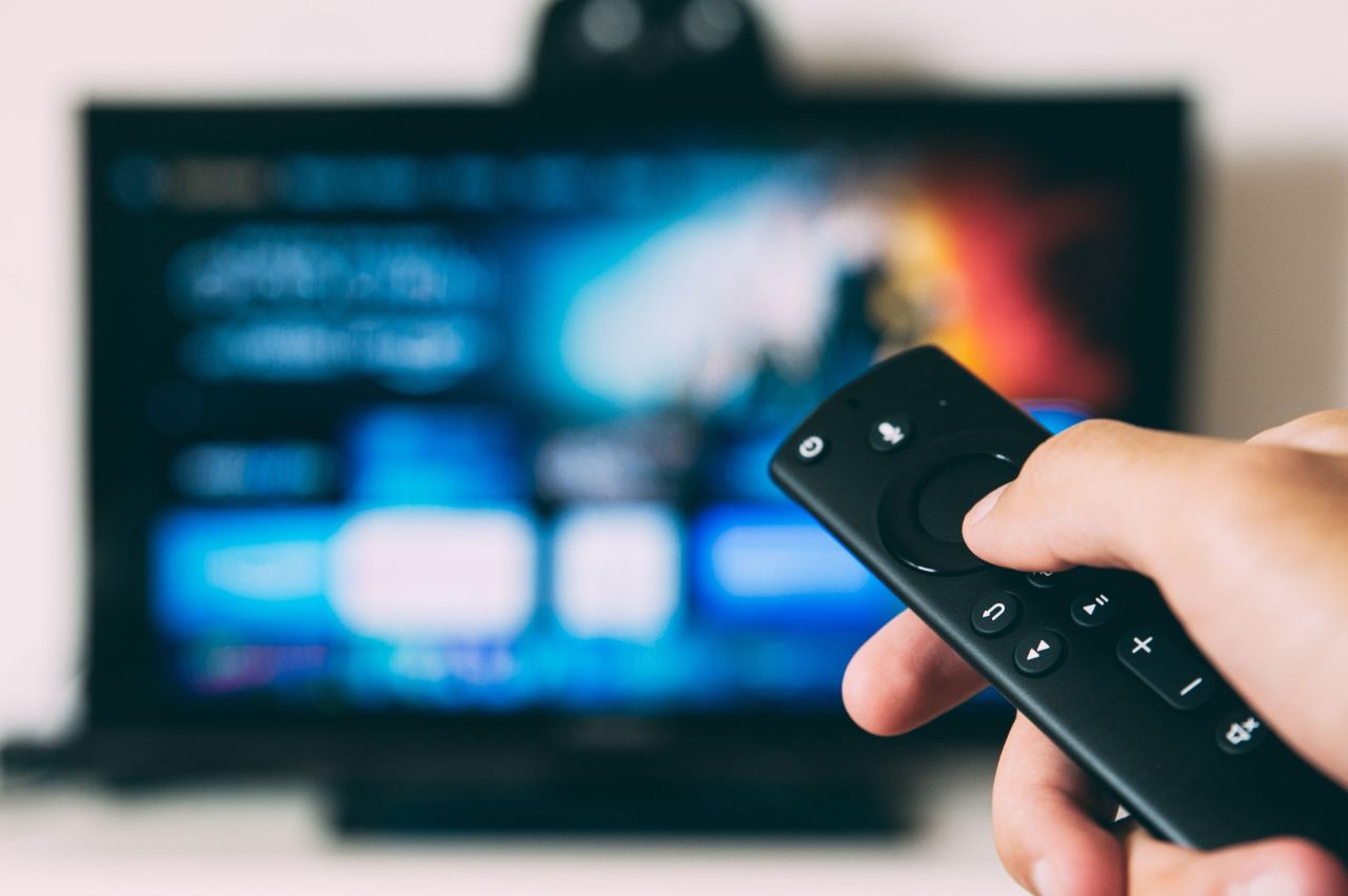 Eine Person hält eine schwarze Fernbedienung in der Hand und richtet sie auf den Fernseher im Hintergrund