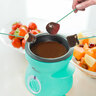 Türkisfarbenes Mini Schoko-Fondue Set mit Teelicht, Schokoladenstückchen, zwei Gabeln und einem Herz-Keks auf einem weißen Untergrund.