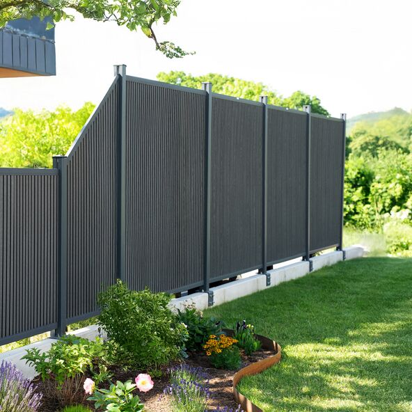 Ein CALLADO Premium Zaunpfosten aus dunklem Material ist vor einem Garten mit Sträuchern und Rasen aufgestellt. Der Pfosten wirkt hochwertig und stabil.