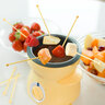 Gelbes Mini Schoko-Fondue Set mit Schokoladenstückchen und kleinen Gabeln, bereit für den Gebrauch auf einer weißen Oberfläche.