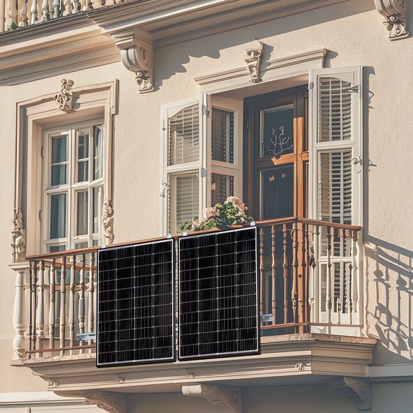 Zwei rechteckige Solarmodule mit schwarzem Rahmen sind auf einem Balkon montiert. Kabel führen zu einem Wechselrichter. Im Hintergrund sind Pflanzen und ein Wohnhaus erkennbar.