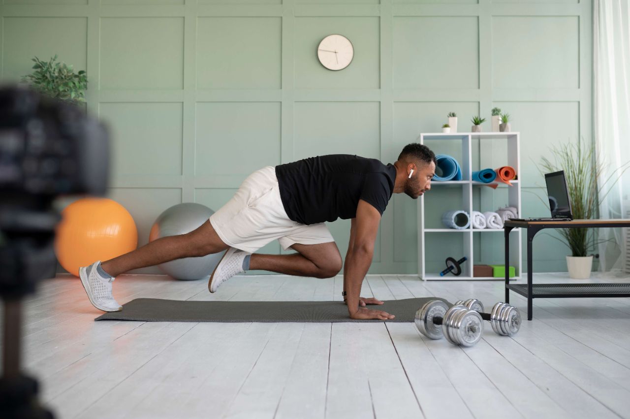 Mann macht Zuhause auf einer Sportmatte ein Workout über seinen Laptop, umgeben von Hanteln und Gymnastikbällen