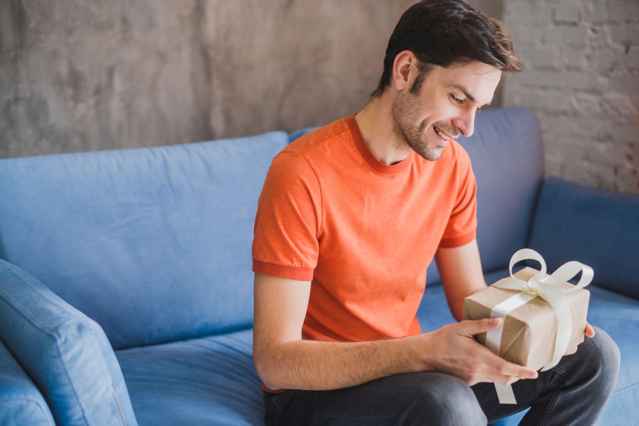 Ein Mann im orangenen T-Shirt sitzt auf einer hellblauen Couch und hält ein Geschenk in beigem Papier mit weißer Schleife