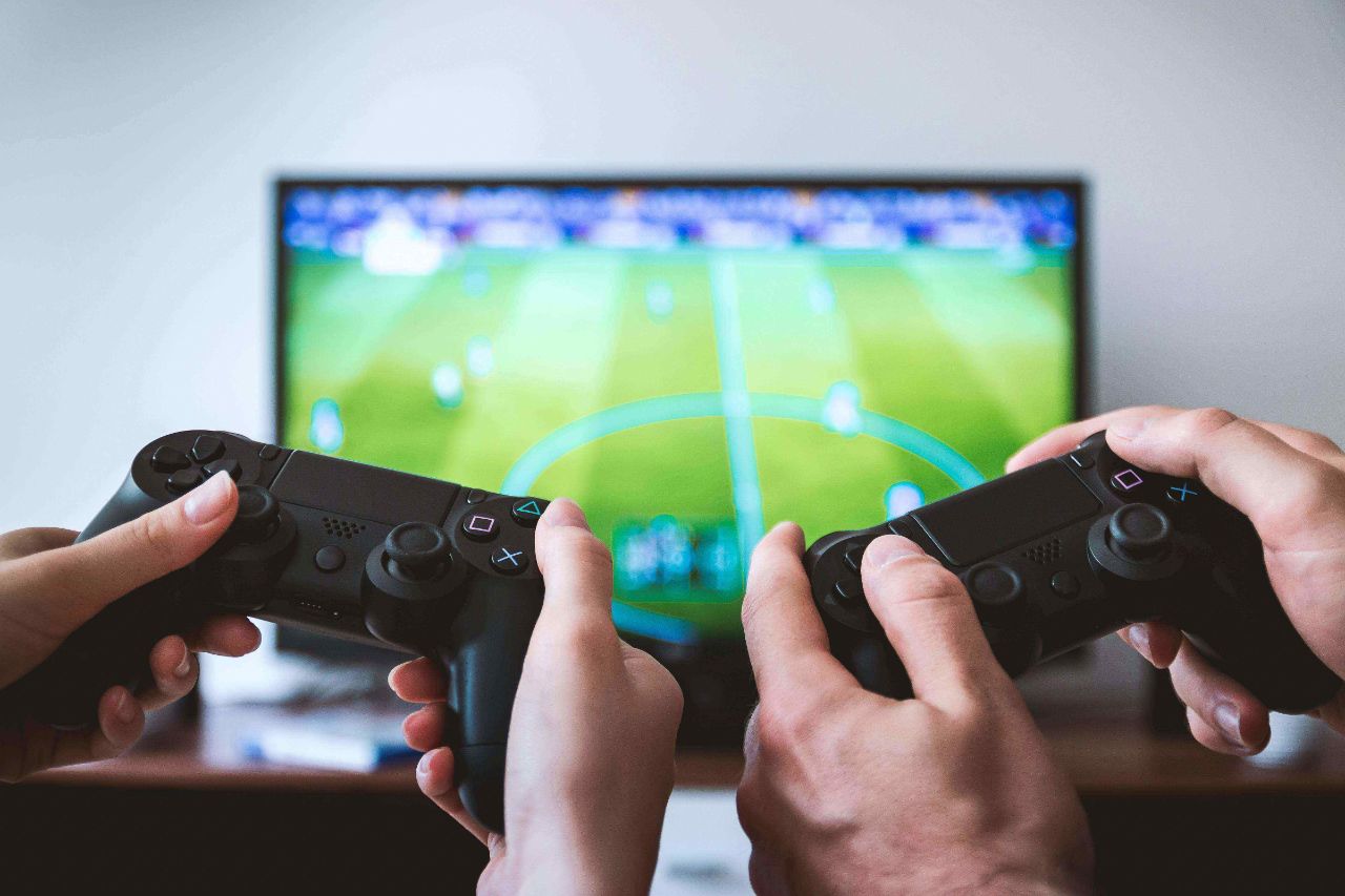 Zwei Personen halten einen schwarzen Controller in der Hand, während im Hintergrund ein Videospiel auf dem Fernseher läuft
