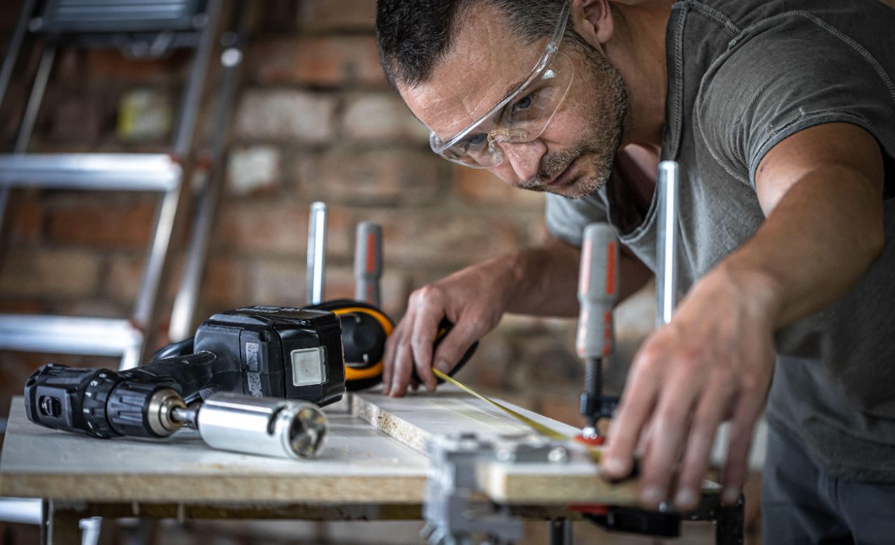 Ein Mann mit Schutzbrille misst in einer Werkstatt ein mit Schraubzwingen befestigtes Holzbrett aus