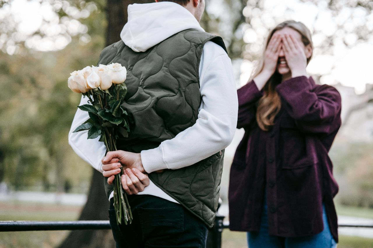Ein Paar steht im Park, die Frau hält ihre Augen zu und der Mann hält ein Bündel weißer Rosen hinter seinem Rücken