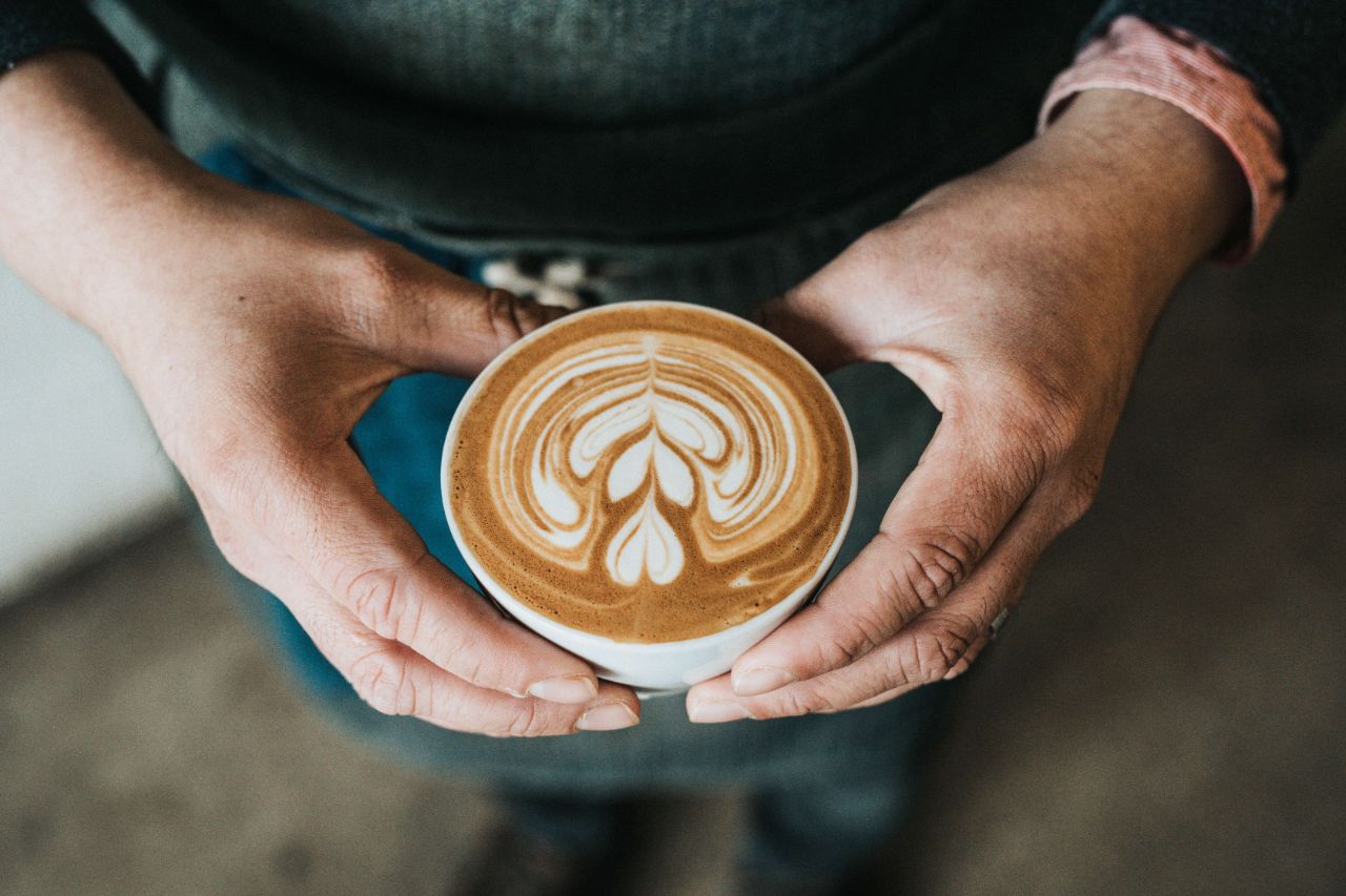 Person hält mit beiden Händen eine weiße Tasse Kaffee, verziert mit einem Blattmuster im Milchschaum