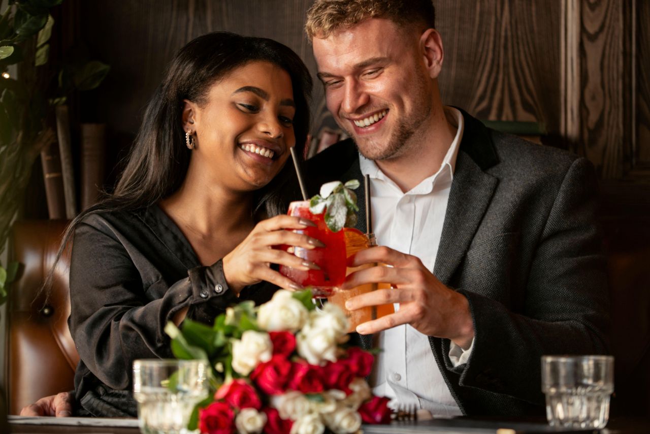 Ein junges Paar sitzt in einem Lokal und stößt mit einem Cocktail an, auf dem Tisch liegen weiße und rote Rosen