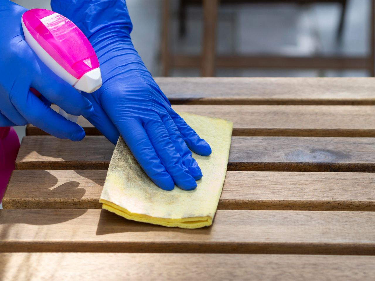 Hände in blauen Handschuhen wischen mit einem gelben Lappen und einer pinken Sprühflasche über eine Holzfläche, sie aus mehreren Sprossen besteht.