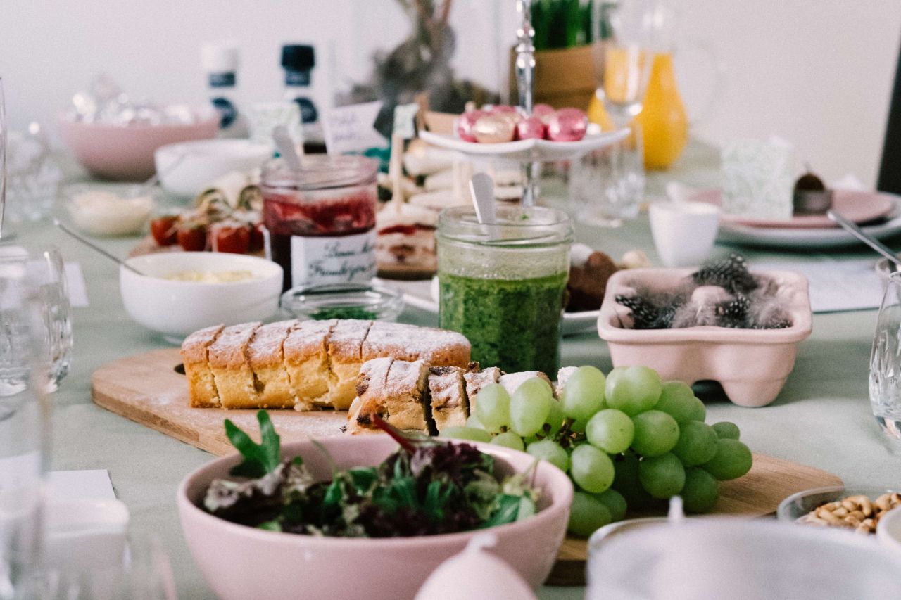 Weiß-rosa gedeckter Tisch mit verschiedenen Speisen wie Salat, Trauben, Kuchen, grünem Pesto und Süßkram auf einer Etagere