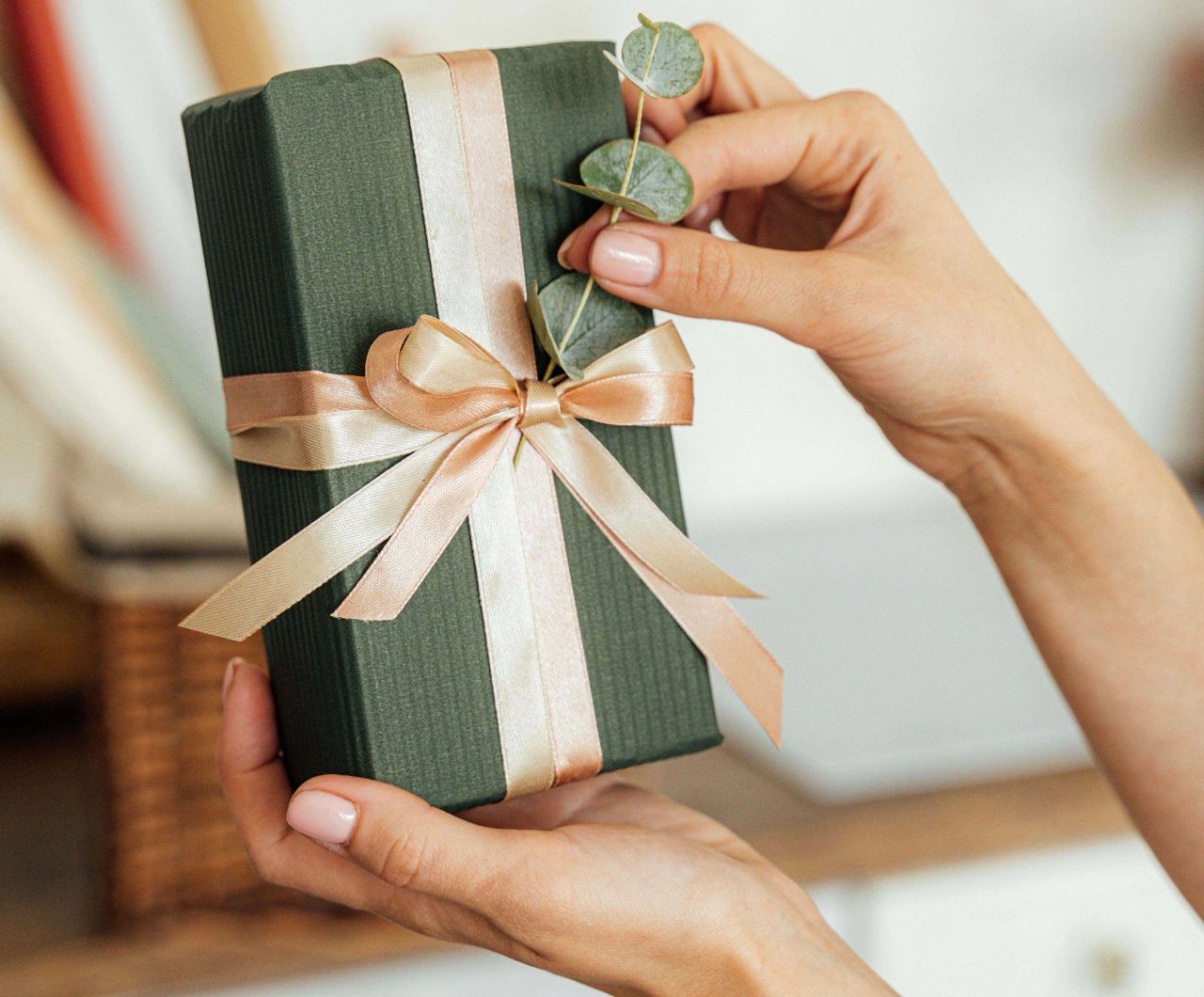 Zwei Hände halten ein Geschenk mit dunkelgrünem Papier und rosa Schleife, verziert mit einem Eukalyptuszweig