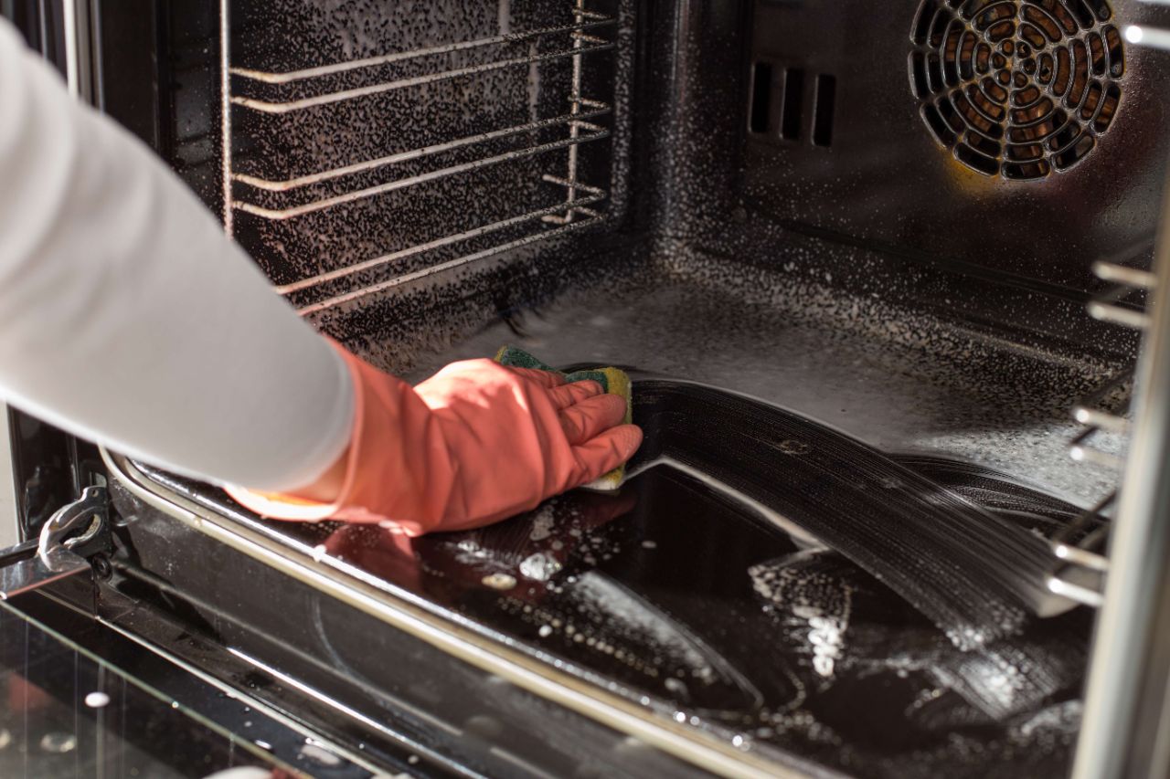 Mit Reiniger eingesprühter Backofen wird von einer Person mit Handschuhen und Schwamm gereinigt