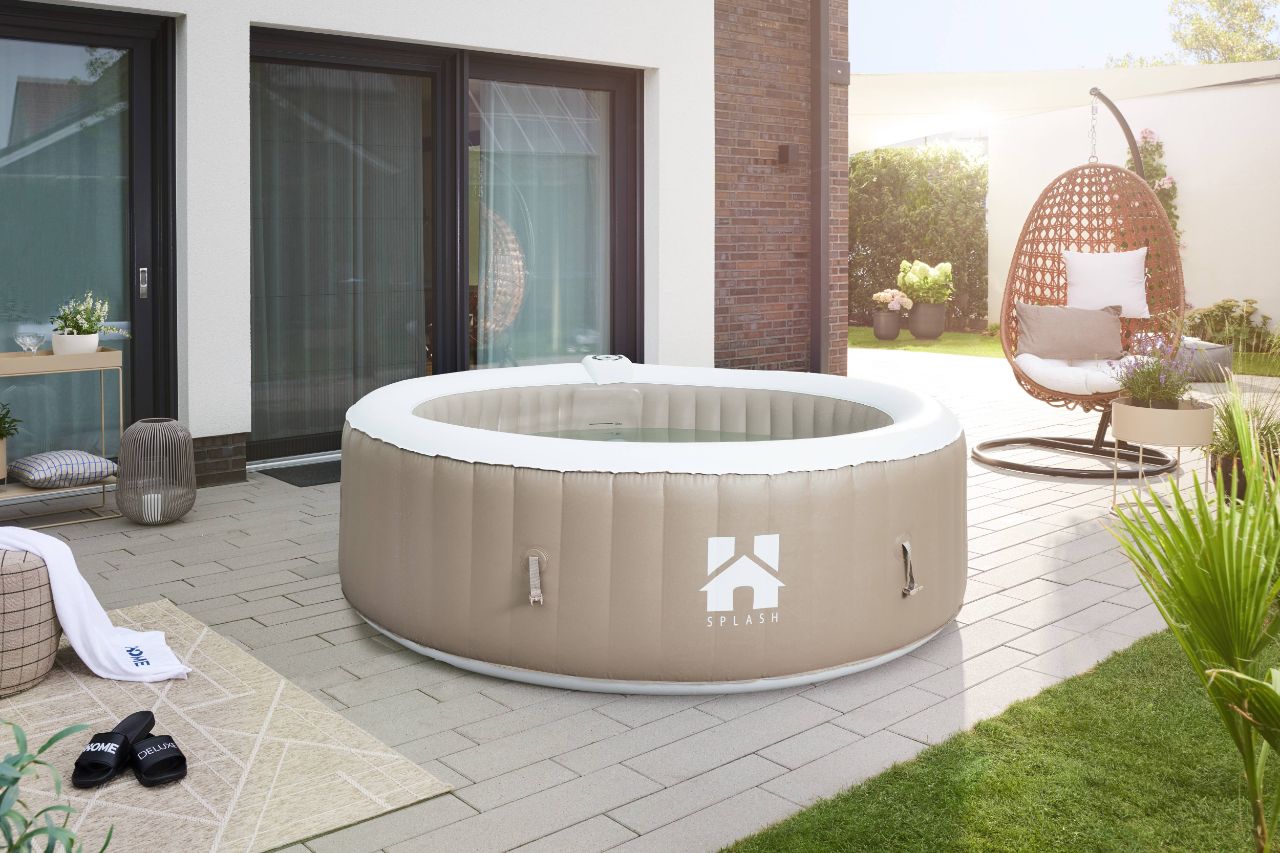 Brauner HOME-DELUXE Whirlpool gefüllt mit Wasser auf einer Terrasse, umgeben von Wiese, Pflanzen und Terrassenmöbeln