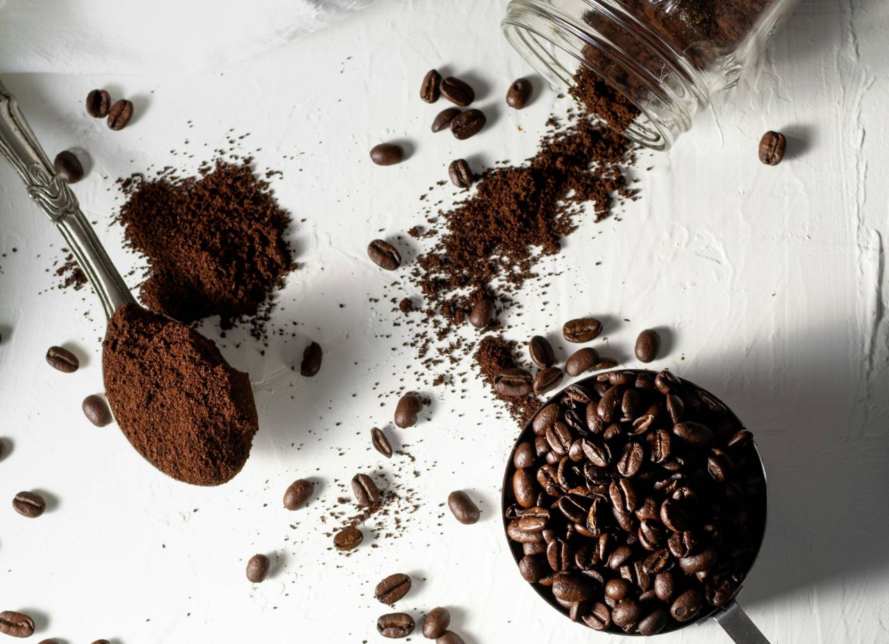 Löffel mit Kaffeepulver, Tasse voller Kaffeebohnen, umgeben von verstreutem Pulver und Bohnen auf weißem Hintergrund