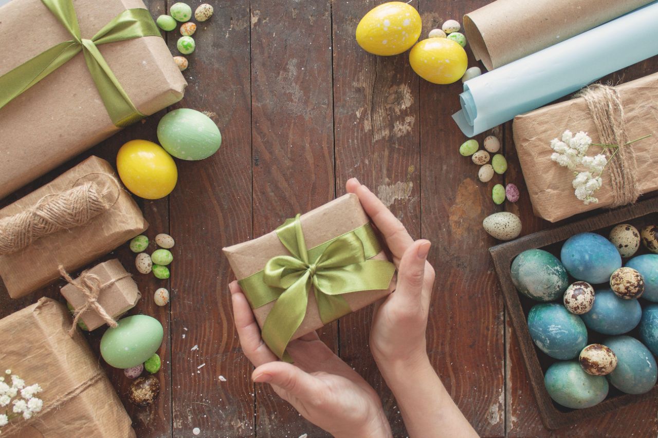 Geschenke und Ostereier auf dunkelbraunem Holztisch, zwei Hände halten ein Geschenk in brauner Verpackung mit grüner Schleife