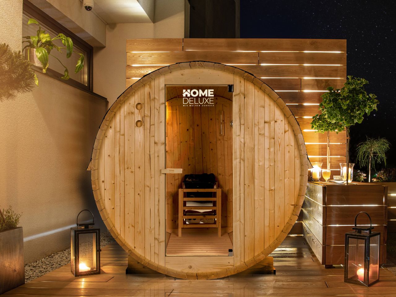 HOME-DELUXE Holzfasssauna auf der Terrasse bei Nacht, umgeben von Pflanzen und sanftem Kerzenschein