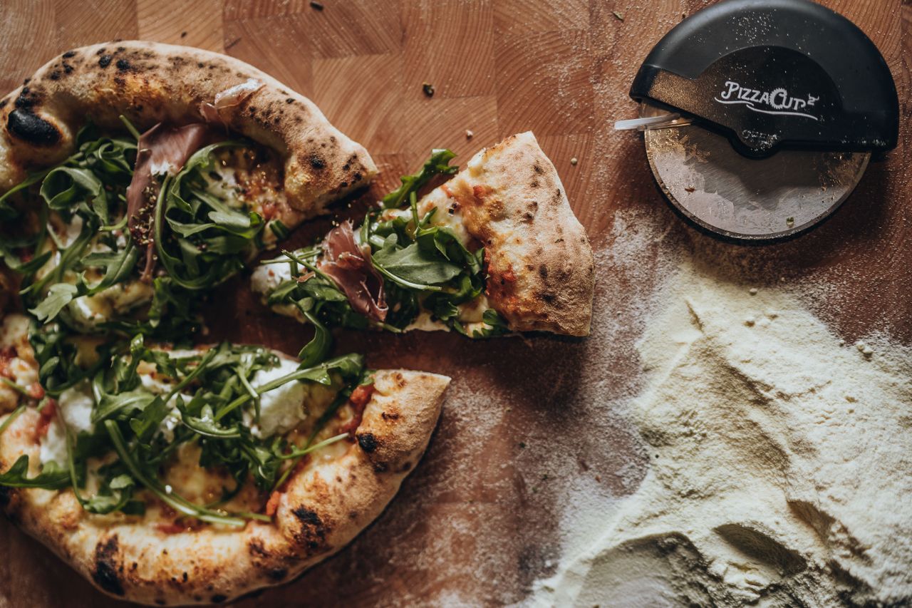 Ein Stück einer Rucola Pizza wurde herausgeschnitten und liegt neben der restlichen Pizza auf einer Holzplatte.