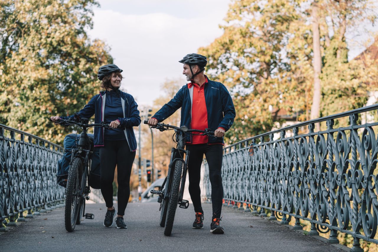 Eine Frau und ein Mann in Fahrradkleidung und Helmen schieben ihr E-Bike über eine Brücke
