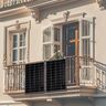 Zwei rechteckige Solarmodule mit schwarzem Rahmen sind auf einem Balkon montiert. Kabel führen zu einem Wechselrichter. Im Hintergrund sind Pflanzen und ein Wohnhaus erkennbar.