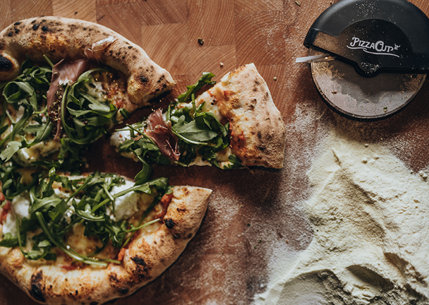 Rucola Pizza auf Holzoberfläche daneben liegt Mehl.
