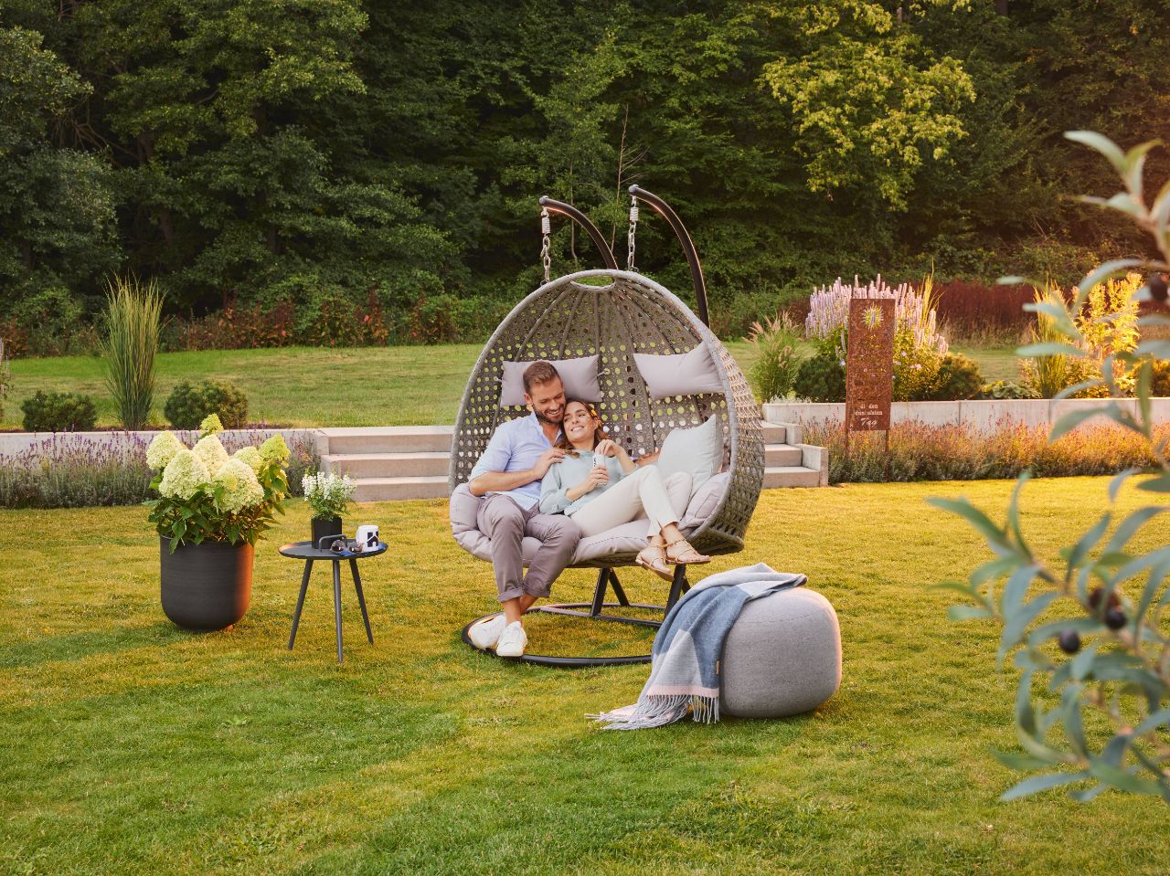 Paar sitzt gemeinsam im Garten in einem Hängesessel im Garten, umgeben von grüner Wiese und Pflanzen